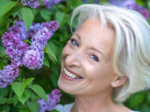 Astrid hat graues Haar, ein freunliches Lächeln und steht im Garten neben einer violetten Blume.